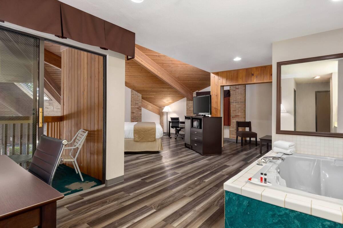 A hotel room with a wooden floor, a large window, a bed, a desk with a computer, a chair, a small table, and a bathtub.