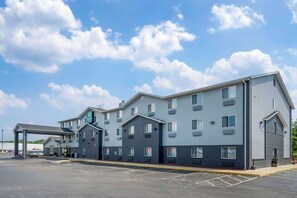 Un hotel de varios pisos con una estación de servicio adosada, que presenta un diseño moderno con una combinación de paredes exteriores grises y azules, múltiples ventanas y un cielo despejado con nubes.