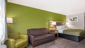 Une chambre d’hôtel avec un mur d’accent vert, un canapé marron, un lit recouvert d’une couverture verte et une table de chevet avec une lampe.