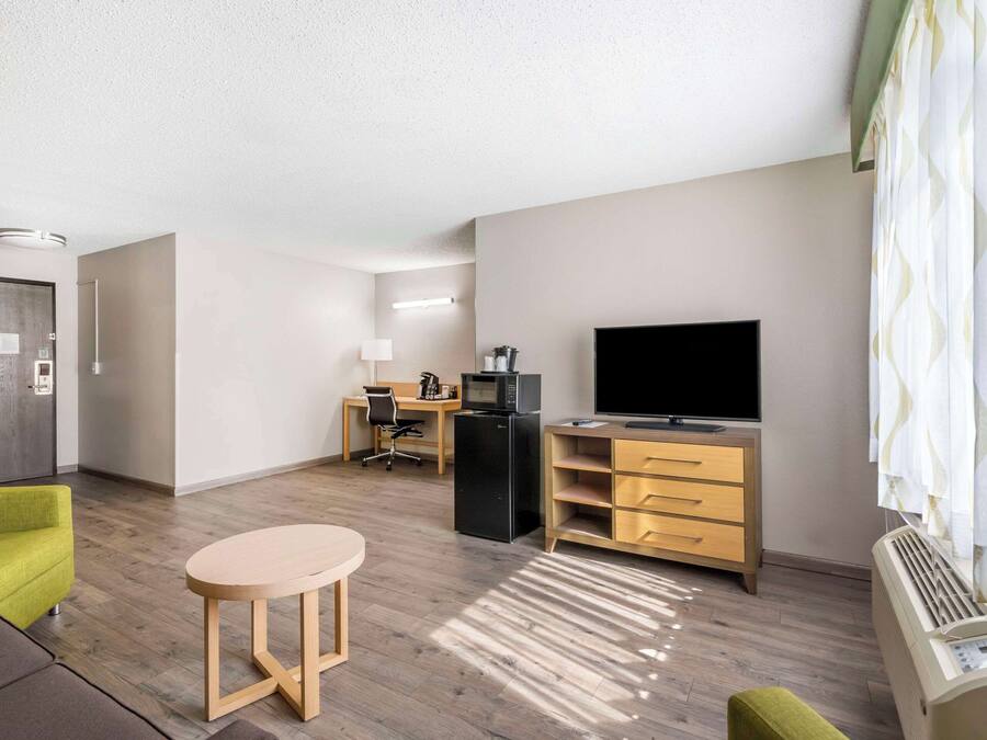 Una habitación de hotel moderna con suelo de madera, un escritorio con computadora y televisor de pantalla plana, una pequeña mesa redonda y un sofá.
