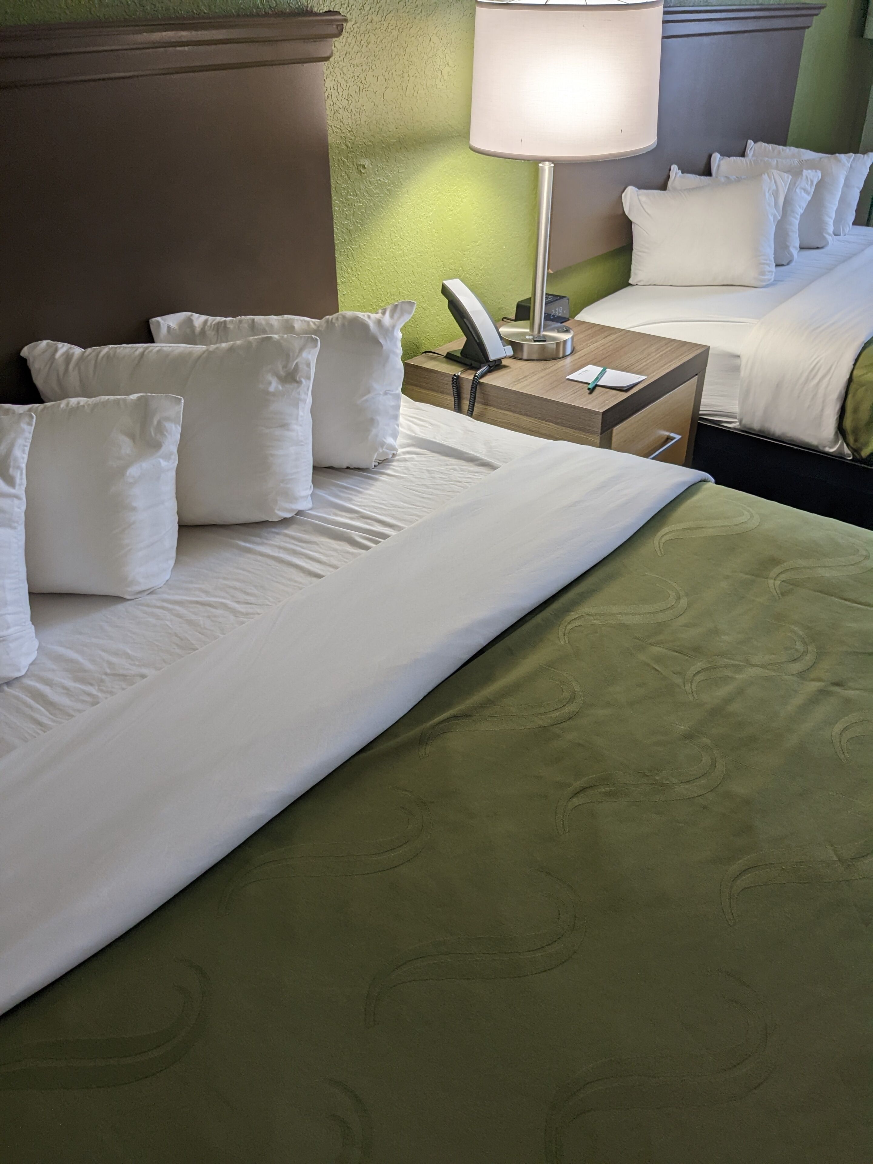 Quarto de hotel com uma cama grande, uma mesa de cabeceira com abajur e um telefone.