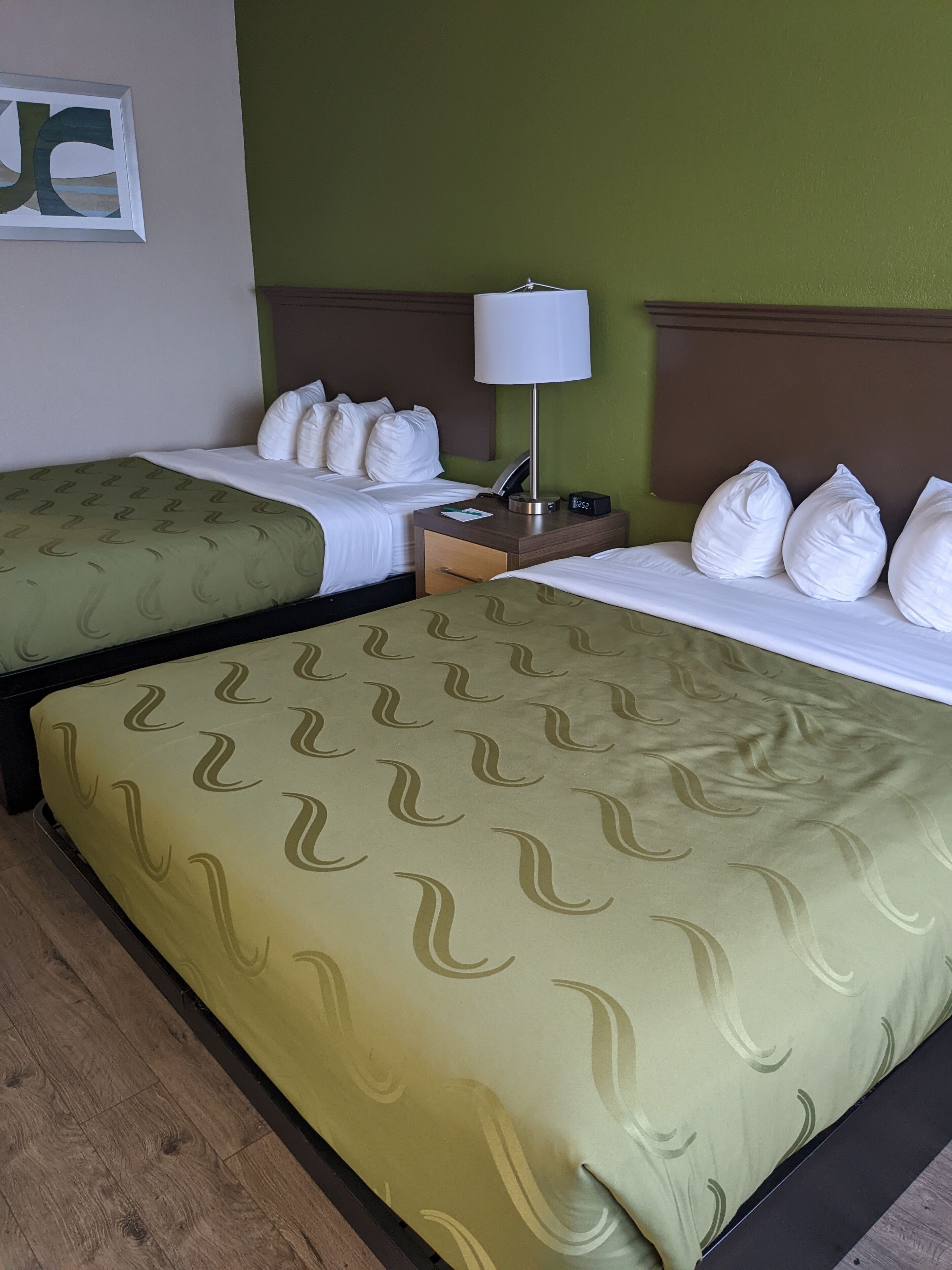 A hotel room with two beds, each with white bedding and pillows, a bedside table with a lamp, and a framed picture on the wall.