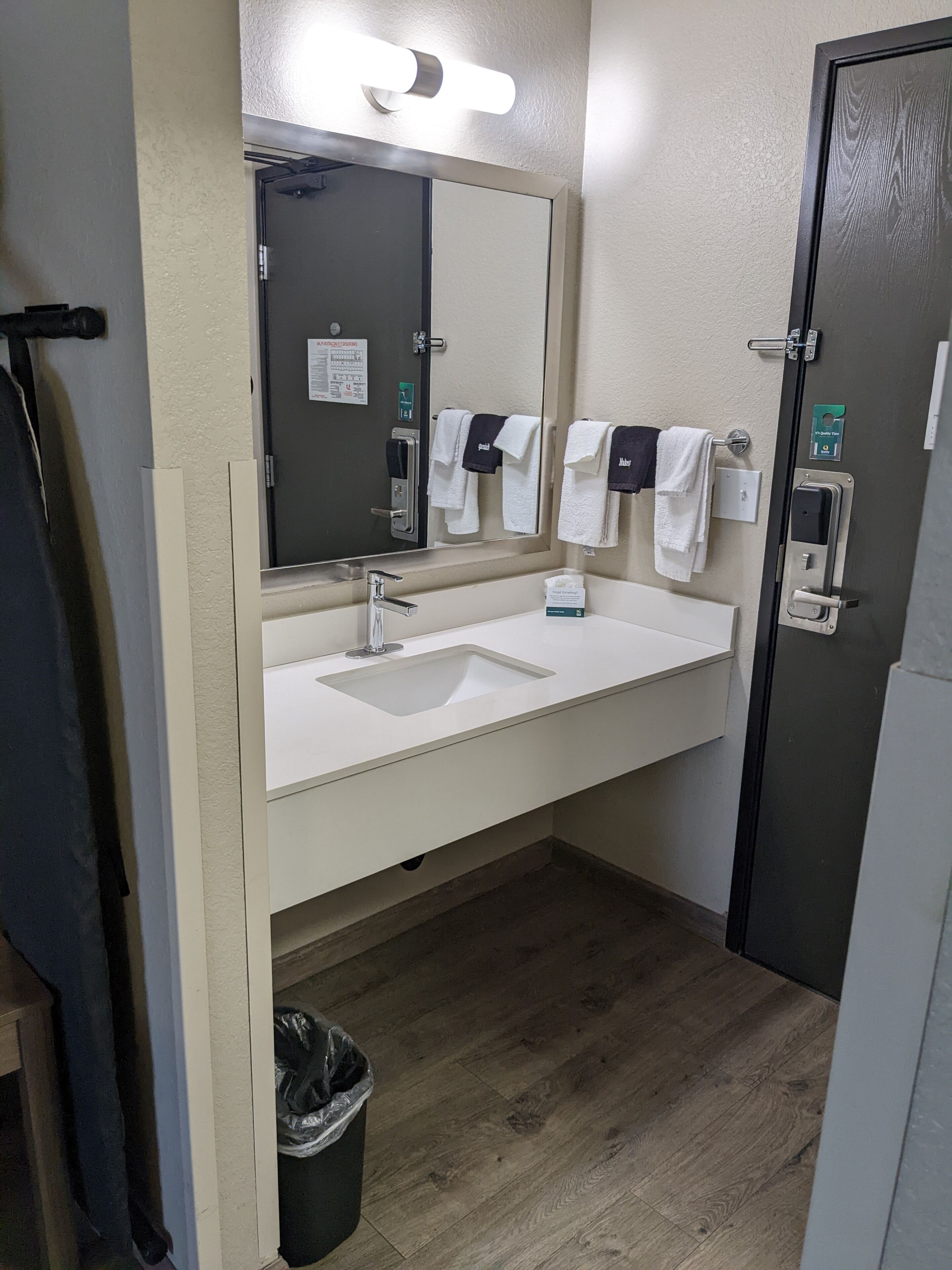 Ein Badezimmer mit einem weißen Waschbecken, einem Spiegel und einem Handtuchhalter. Ein Mülleimer und eine Tür mit Griff sind ebenfalls vorhanden.