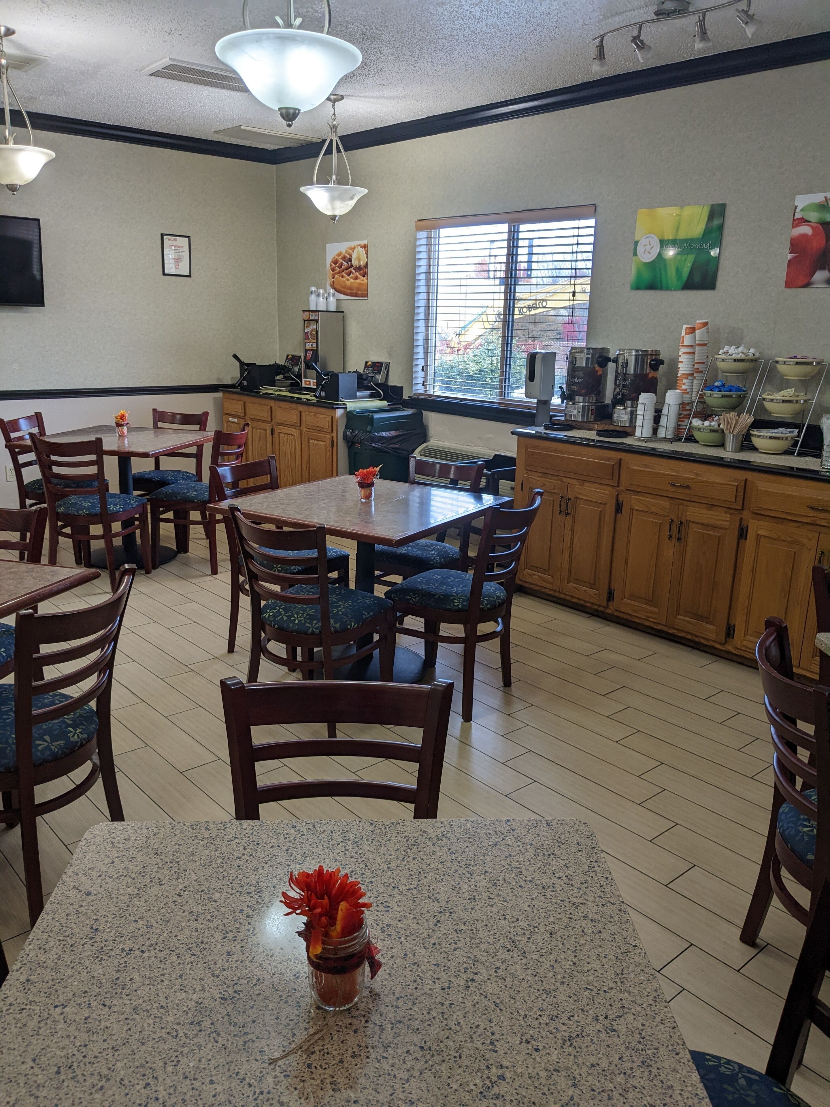 A restaurant with wooden tables and chairs, a kitchen area, and a window with blinds.