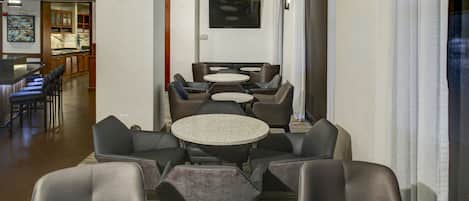 A hotel lobby with a seating area featuring round tables and chairs, a television mounted on the wall, and a bar area in the background.