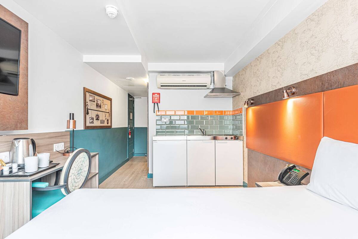 A compact hotel room with a kitchenette, a bed, a desk with a chair, and a telephone.