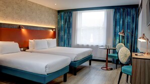A hotel room with two beds, a desk, a chair, and a window with curtains.