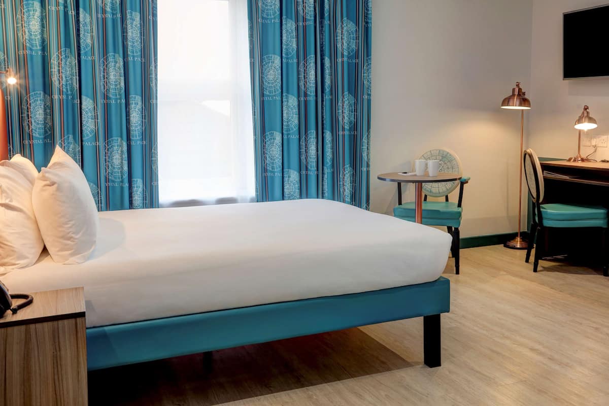 A hotel room with a bed, a desk, a chair, a lamp, and curtains.