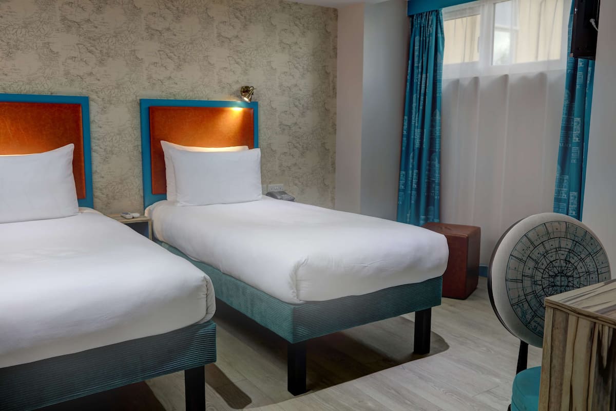 A hotel room with two single beds, a wooden headboard, a bedside table with a phone, a small round table with a decorative item, and a wall-mounted fan.