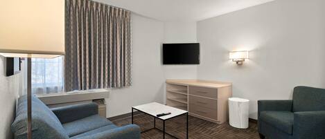 Habitación de hotel con sofá, una mesita, televisor montado en la pared, lámpara y ventana con cortinas.