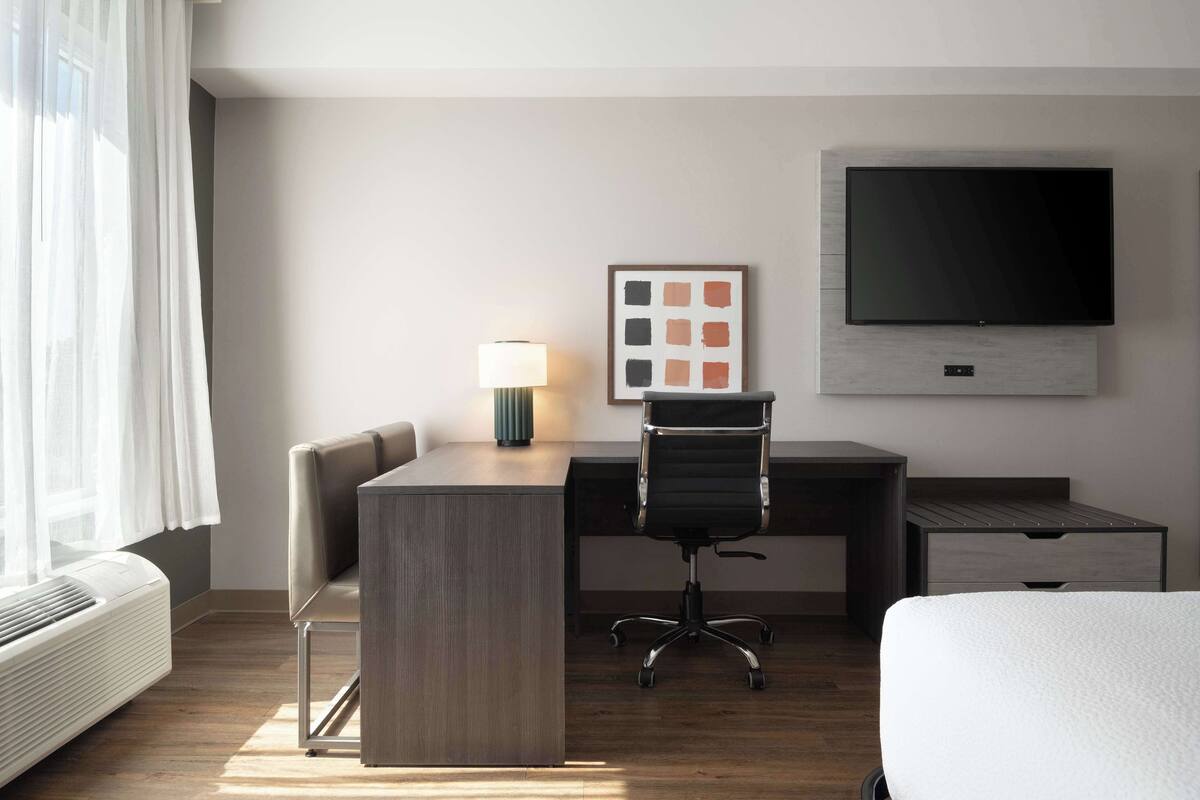 A hotel room with a bed, a desk, a chair, a TV, a lamp, and a window with curtains.