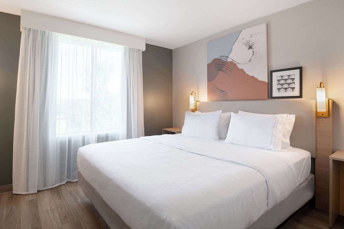 A hotel room with a large bed, a window with sheer curtains, a wall-mounted artwork, and two bedside lamps.