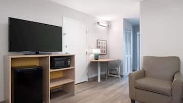 A compact living space with a flat-screen TV on a wooden stand, a small refrigerator, a microwave, a beige armchair, a small table with a lamp, and a framed picture on the wall.