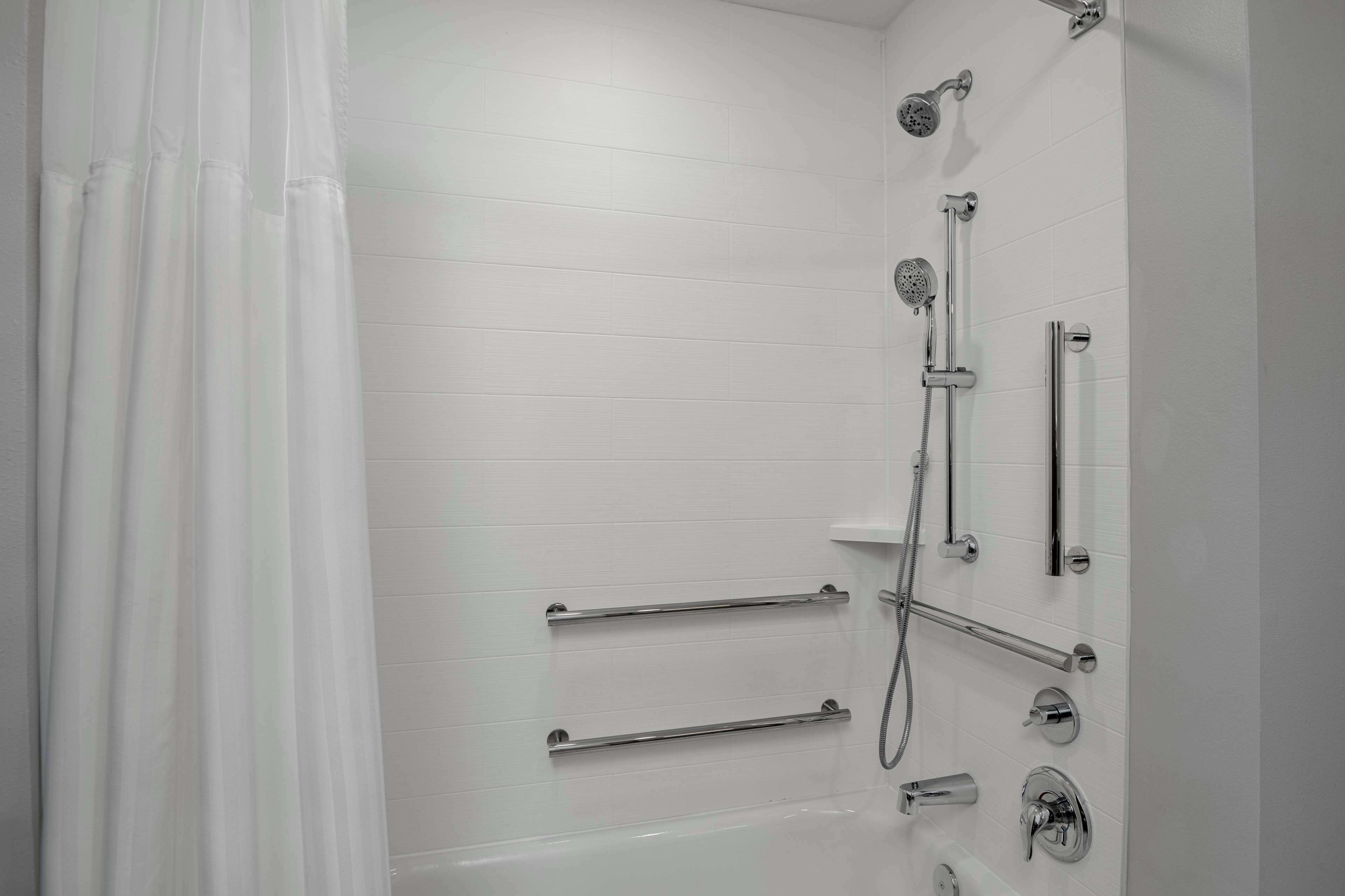 A modern bathroom with white tiled walls, a bathtub, a shower with multiple fixtures, and grab bars.