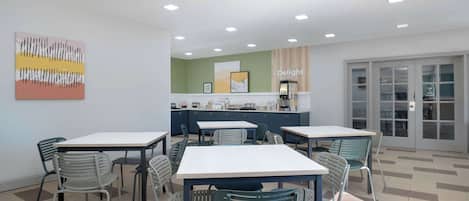 A modern dining area with tables and chairs, a kitchen in the background, and a large window with a view.