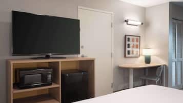 Habitación de hotel con cama, televisión sobre un soporte de madera, microondas, refrigerador pequeño, escritorio con silla y puerta.