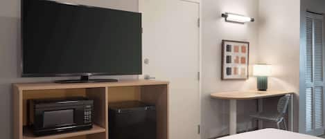 Une chambre d’hôtel comprenant un lit, un téléviseur posé sur un meuble en bois, un four à micro-ondes, un mini-réfrigérateur, un bureau avec une chaise et une porte.