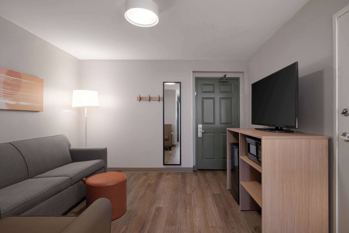 A modern living room with a grey sofa, a round orange ottoman, a floor lamp, a flat-screen TV on a wooden stand, and a mirror on the wall.