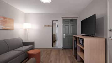 A modern living room with a grey sofa, a round orange ottoman, a floor lamp, a flat-screen TV on a wooden stand, and a mirror on the wall.
