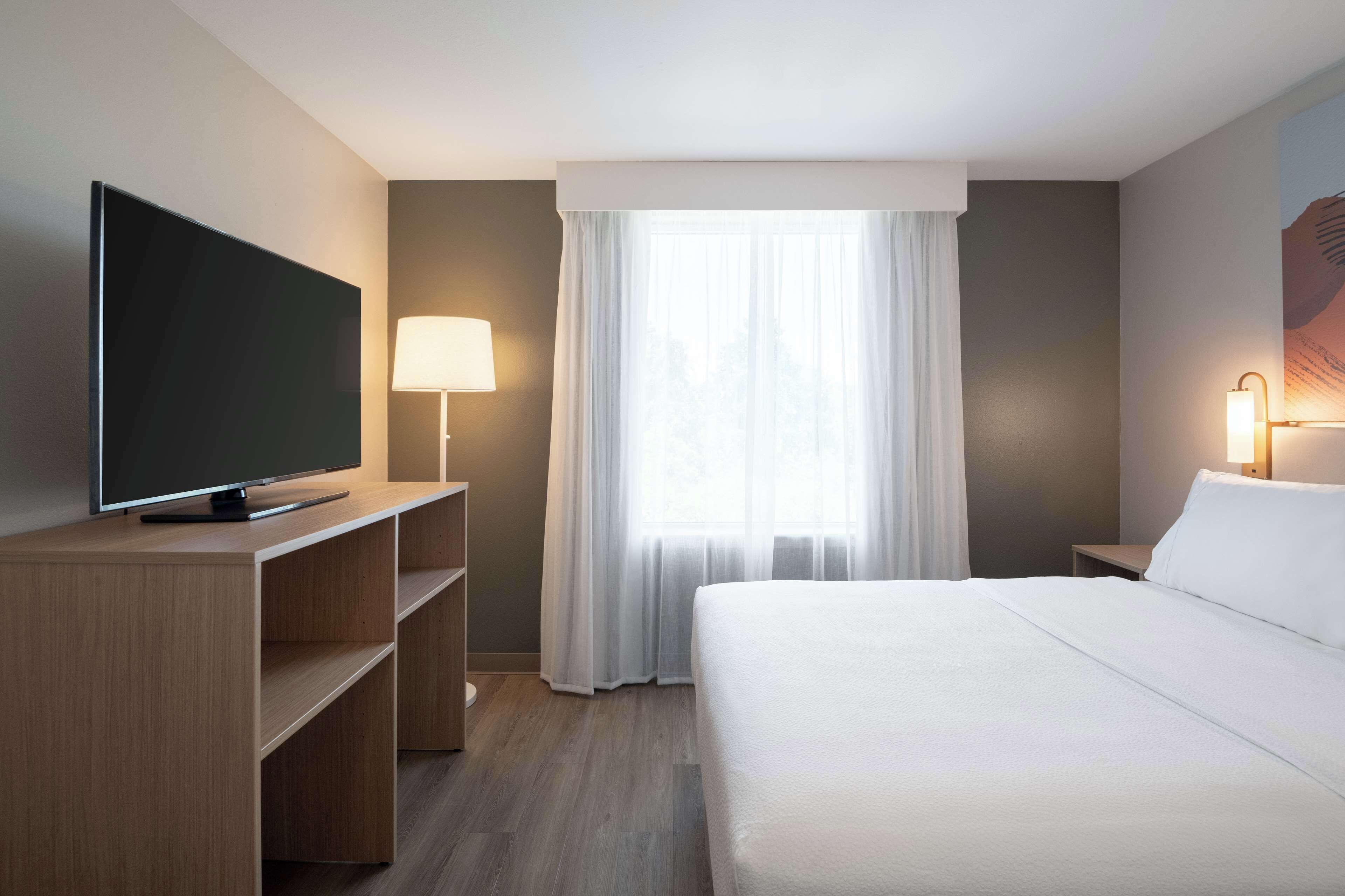 A hotel room with a flat-screen TV on a wooden stand, a bedside lamp, a large bed with white linens, and a window with sheer curtains.