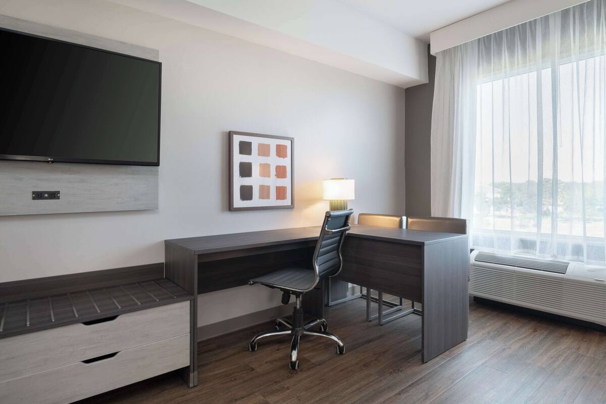 A modern hotel room with a large flat-screen TV, a desk with a chair, a framed artwork, a lamp, and a large window with curtains.