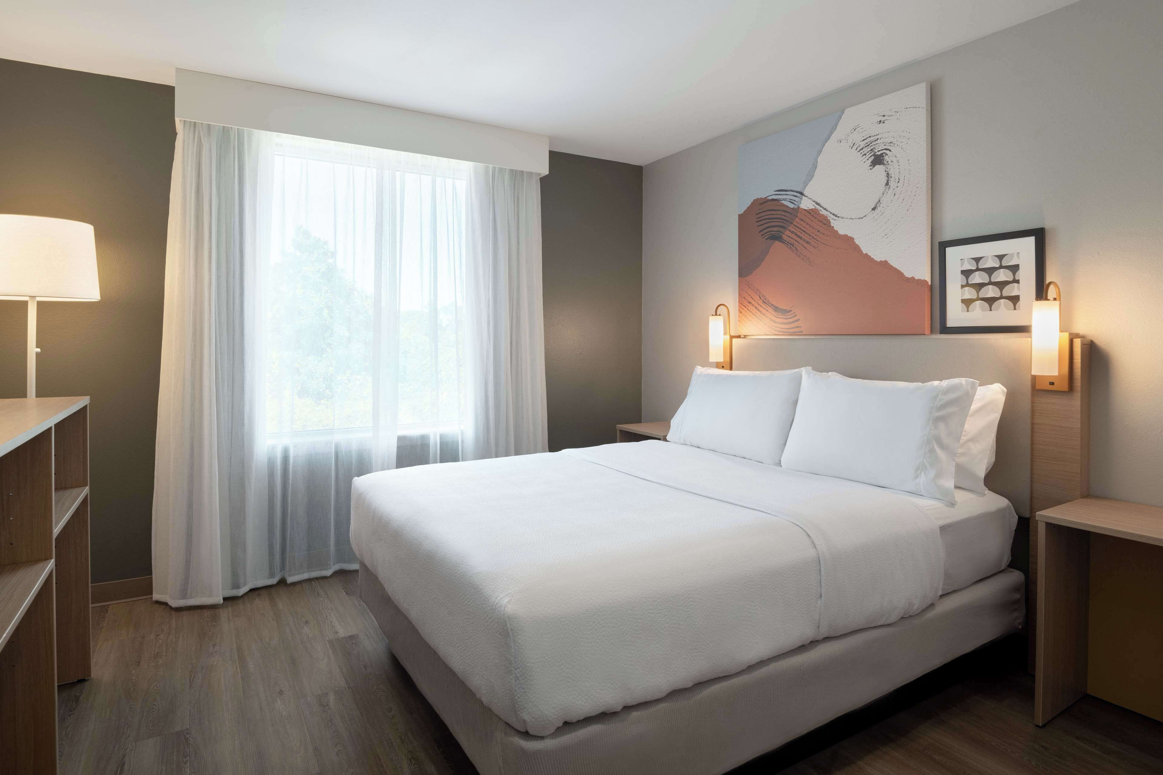 A hotel room with a large bed, a bedside table, a lamp, a window with sheer curtains, and artwork on the wall.