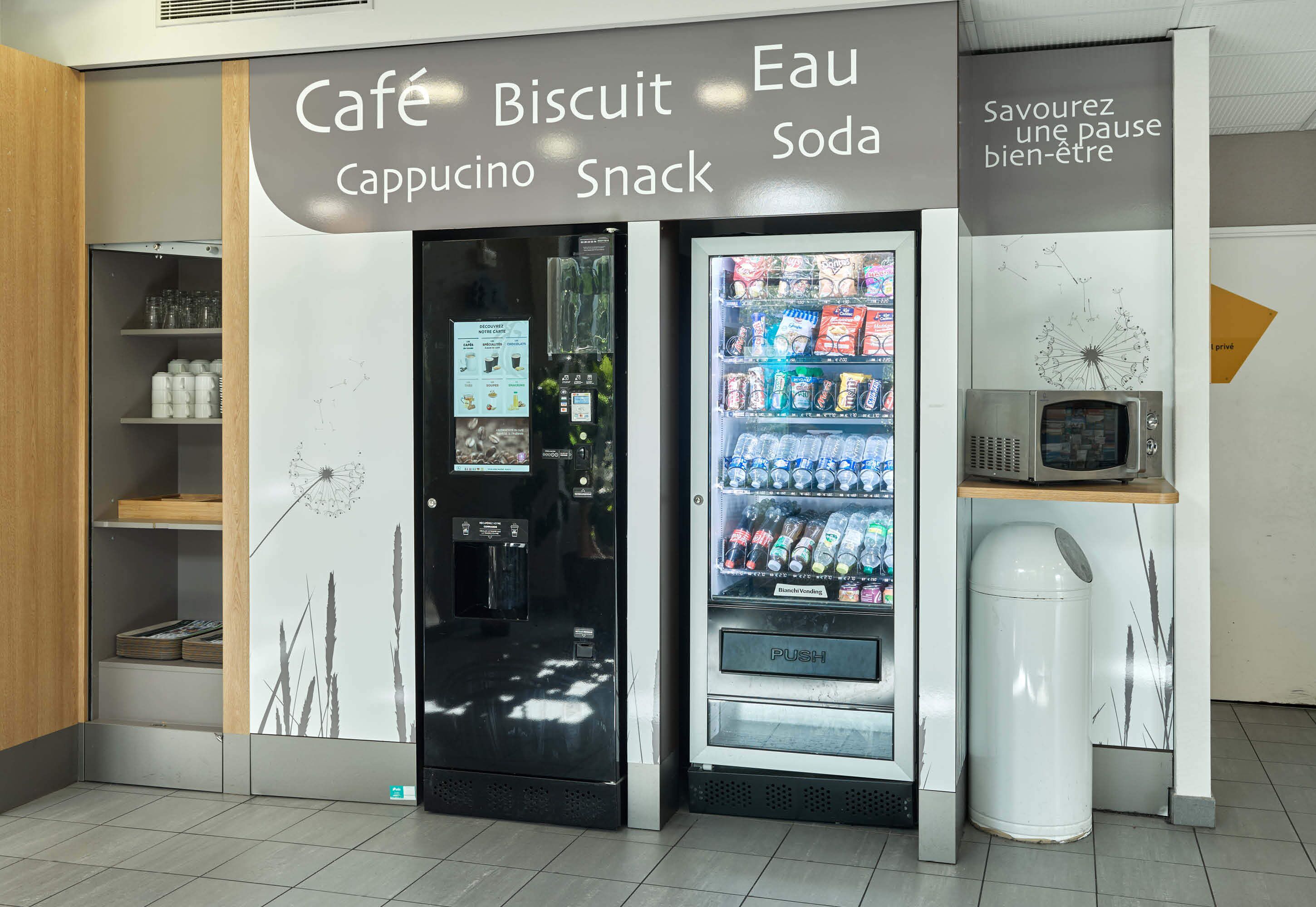 A café area with a beverage vending machine and a snack vending machine, shelves with cups and a dandelion decoration.