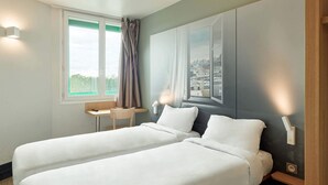 A hotel room with two beds, a small desk, a chair, a window with a view of a cityscape, and a wall-mounted light fixture. - B&B HOTEL Orly Rungis Aéroport (Rungis)