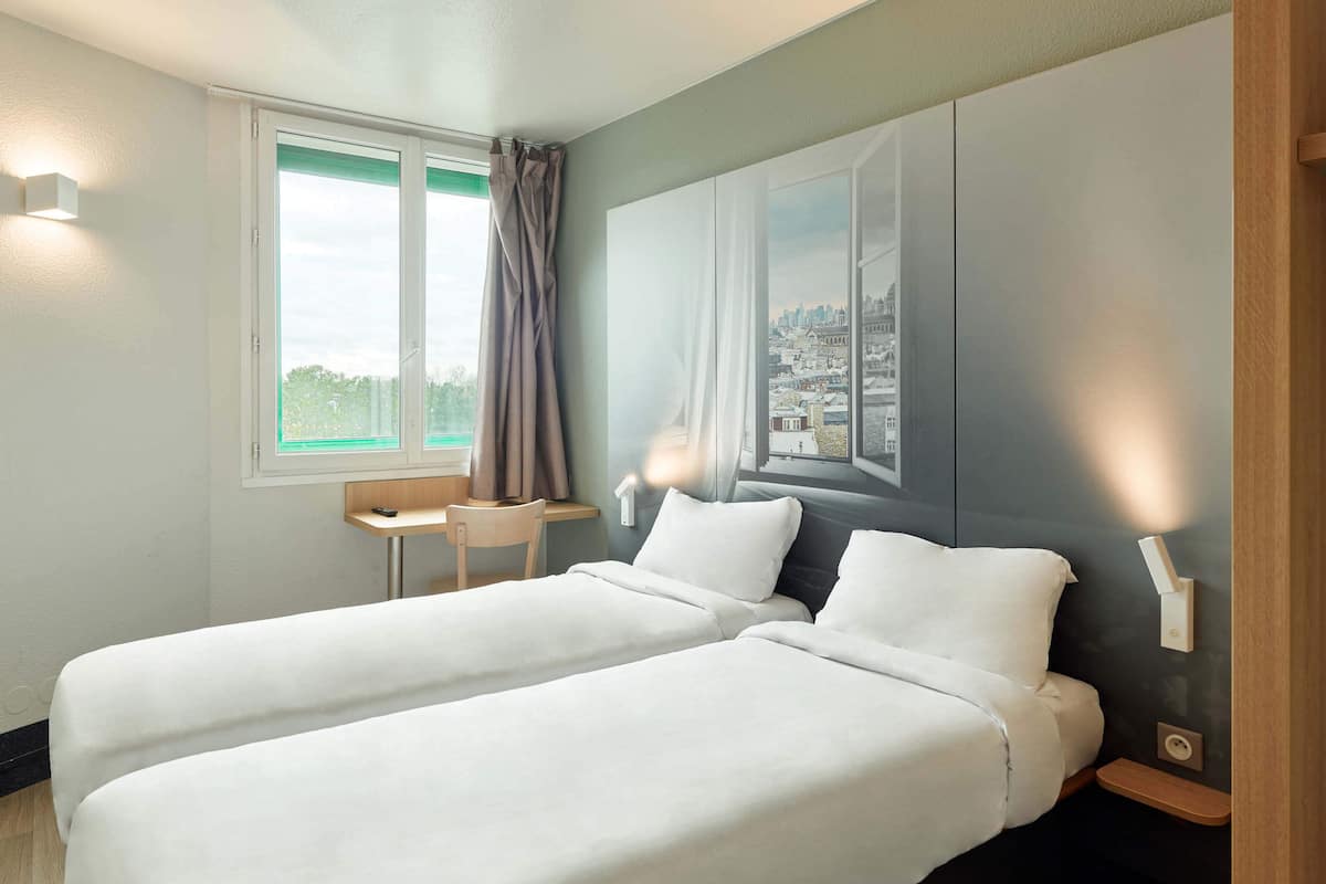 Habitación de hotel con dos camas, un escritorio pequeño, una silla, un ventanal con vistas a un paisaje urbano y una lámpara fijada a la pared.
