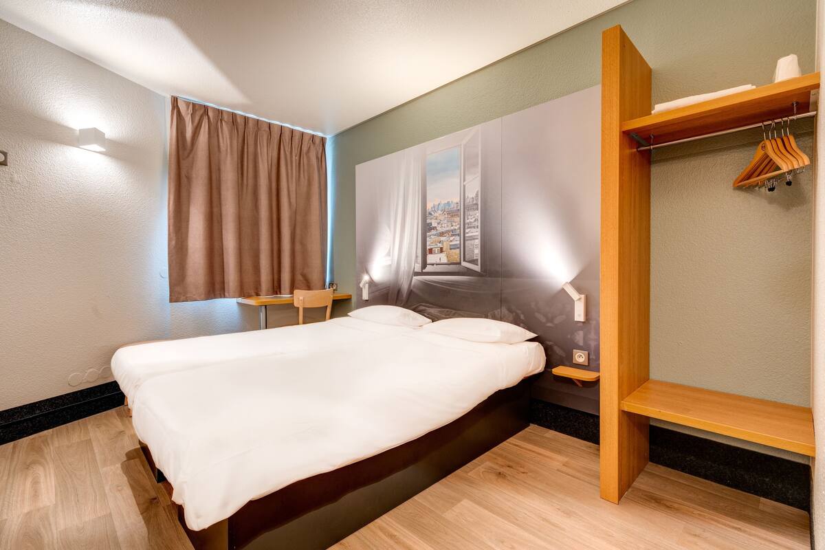 Una habitación de hotel con cama, una ventana con cortinas, un estante de madera con perchas y una mesita.