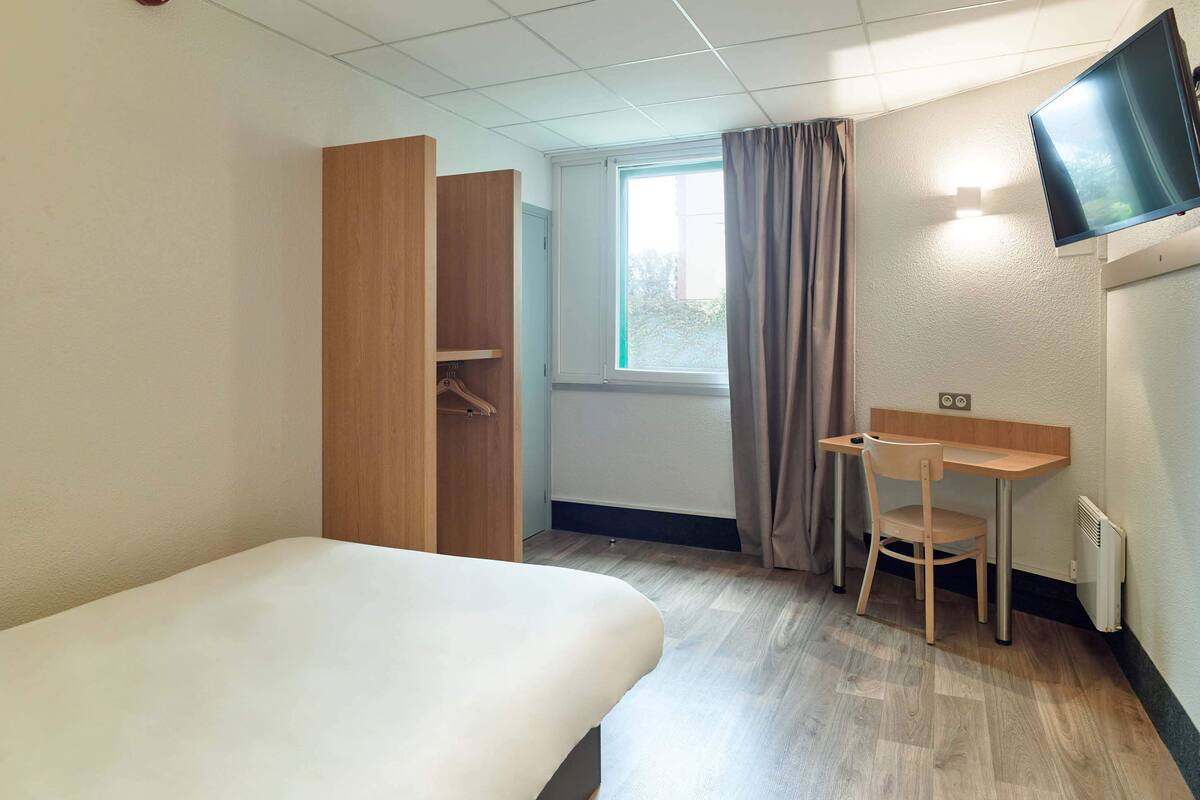 Habitación de hotel con cama, escritorio, silla, televisor, ventana con cortinas y armario.