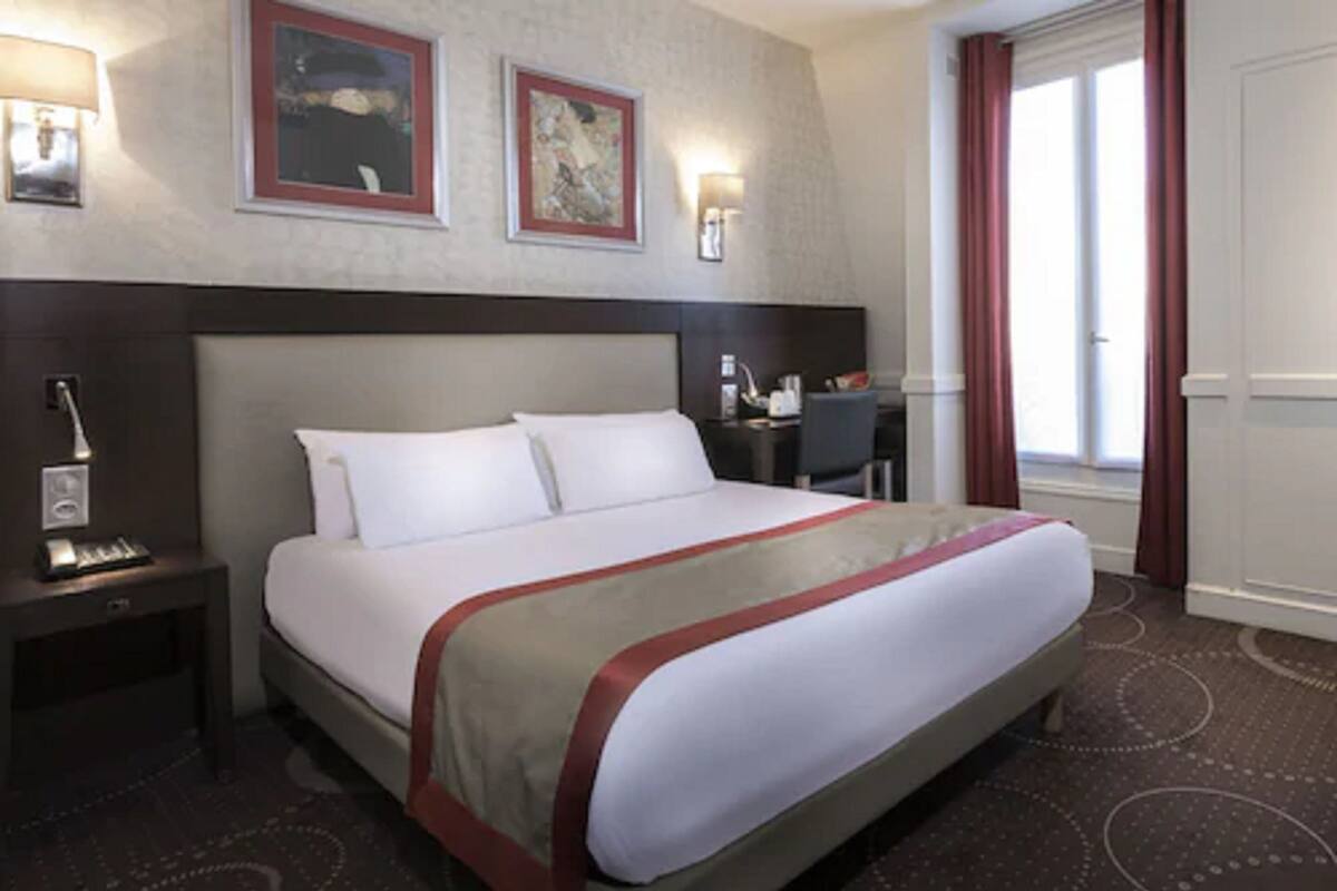 Une chambre d’hôtel avec un grand lit, une table de chevet, un bureau et une fenêtre avec des rideaux.
