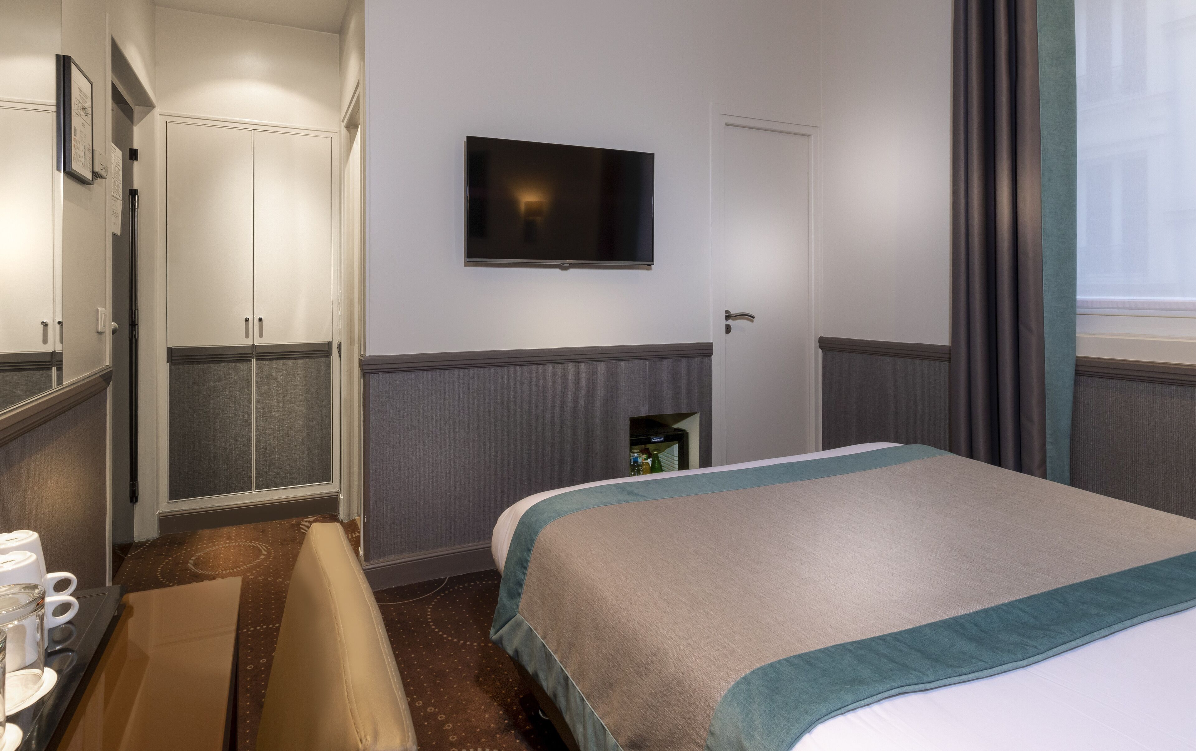 A hotel room with a bed, a television mounted on the wall, a wardrobe, and a small cabinet.
