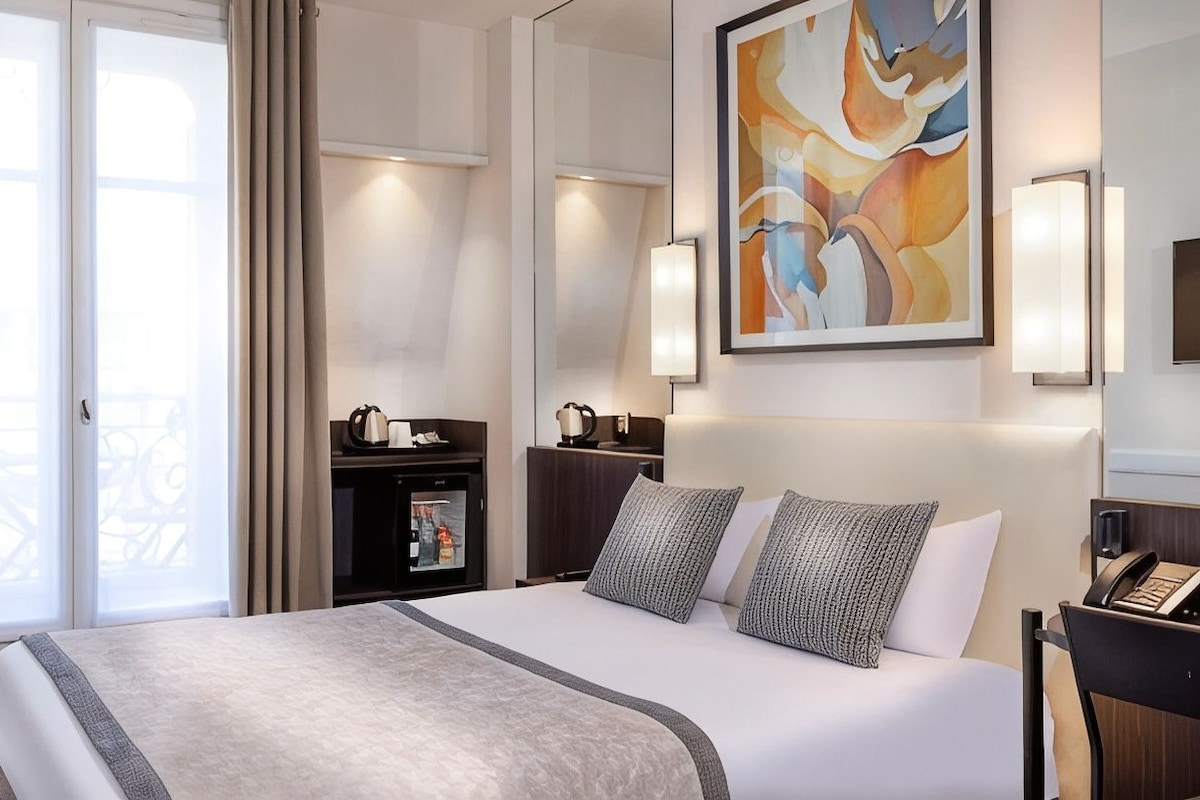 A hotel room with a large bed, a desk with a telephone, a minibar, and a large abstract painting on the wall.