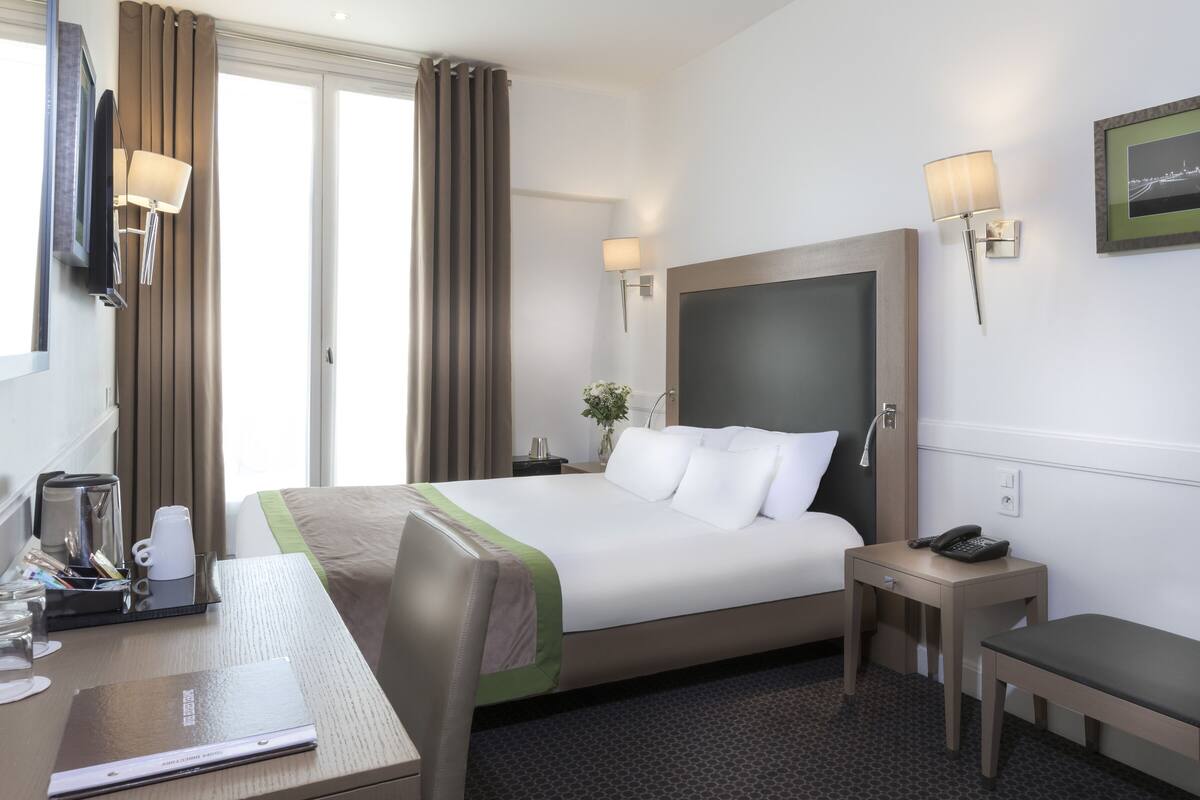 Une chambre d’hôtel avec un grand lit, un bureau avec une chaise, une petite table, un téléphone et un cadre avec une image accroché au mur.
