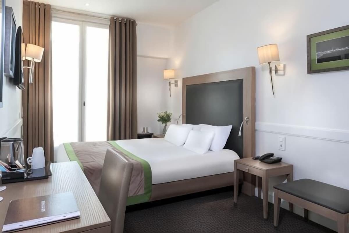 A hotel room with a bed, bedside table, lamp, and a desk with a chair.