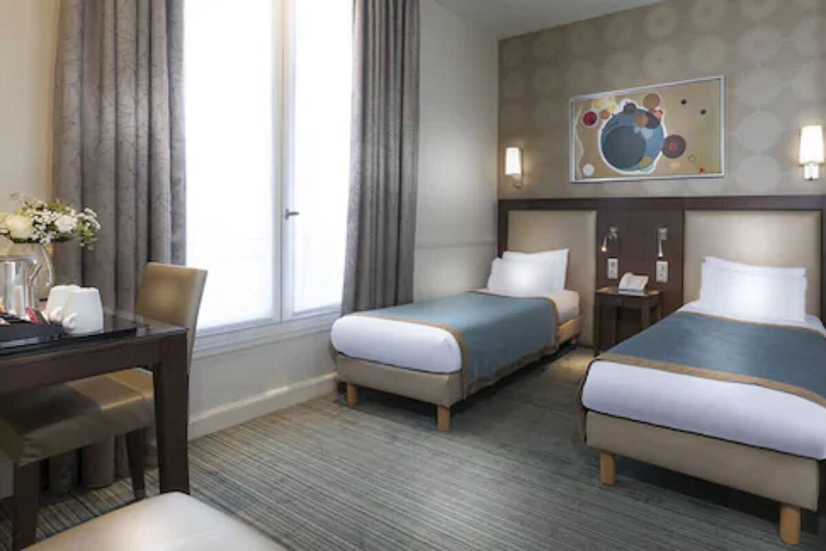 Une chambre d’hôtel avec deux lits, un bureau et une chaise. Il y a une fenêtre avec des rideaux, un tableau au mur et un vase avec des fleurs sur le bureau.
