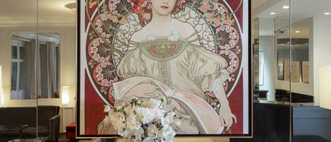A framed artwork of a woman in a detailed dress, a vase of white flowers, and a marble table in a modern room with a large mirror and a hanging light fixture.