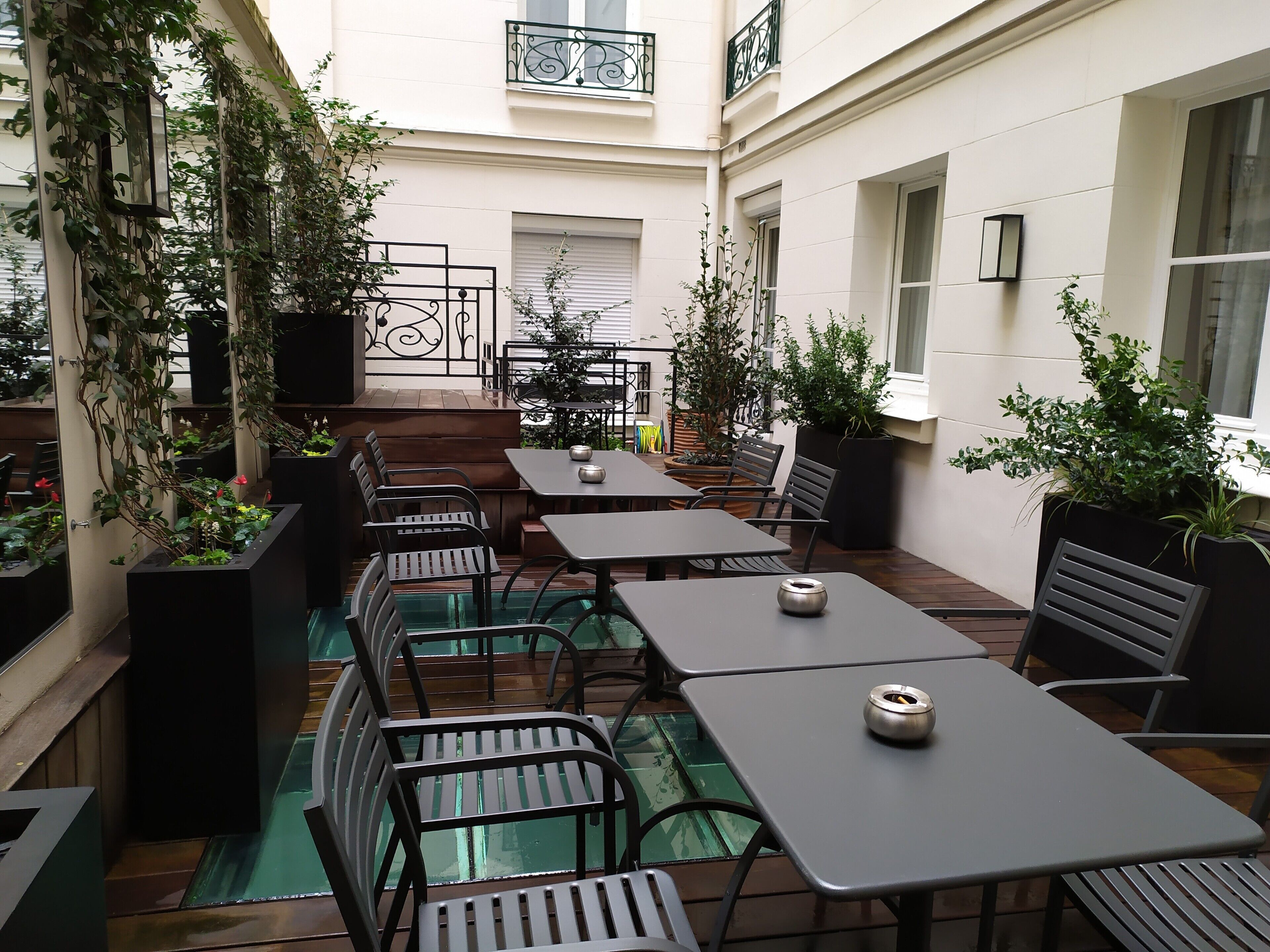 An outdoor café with tables and chairs, surrounded by potted plants and a glass enclosure.