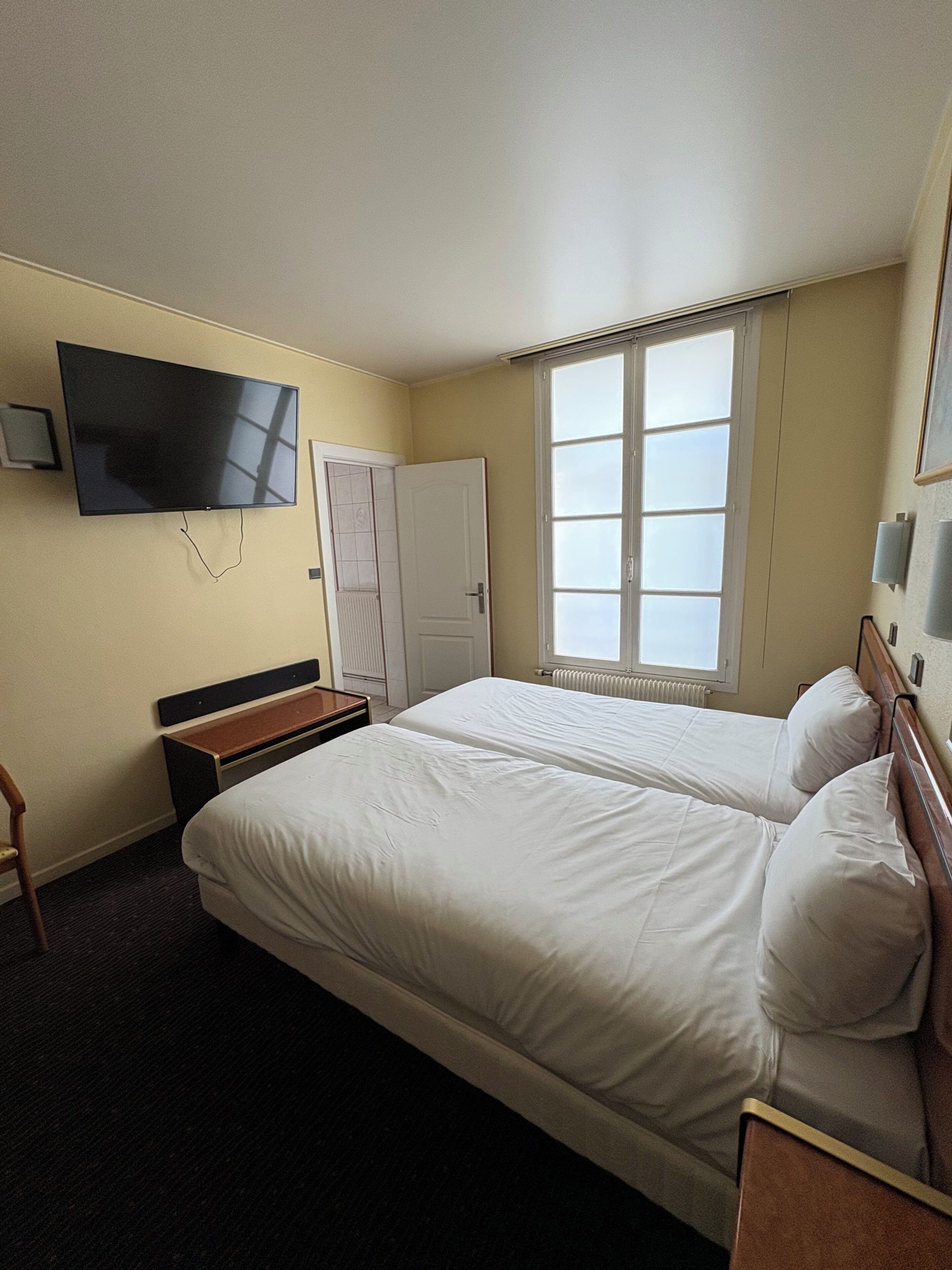 A hotel room with two beds, a TV, a window with blinds, a door, and a small table.