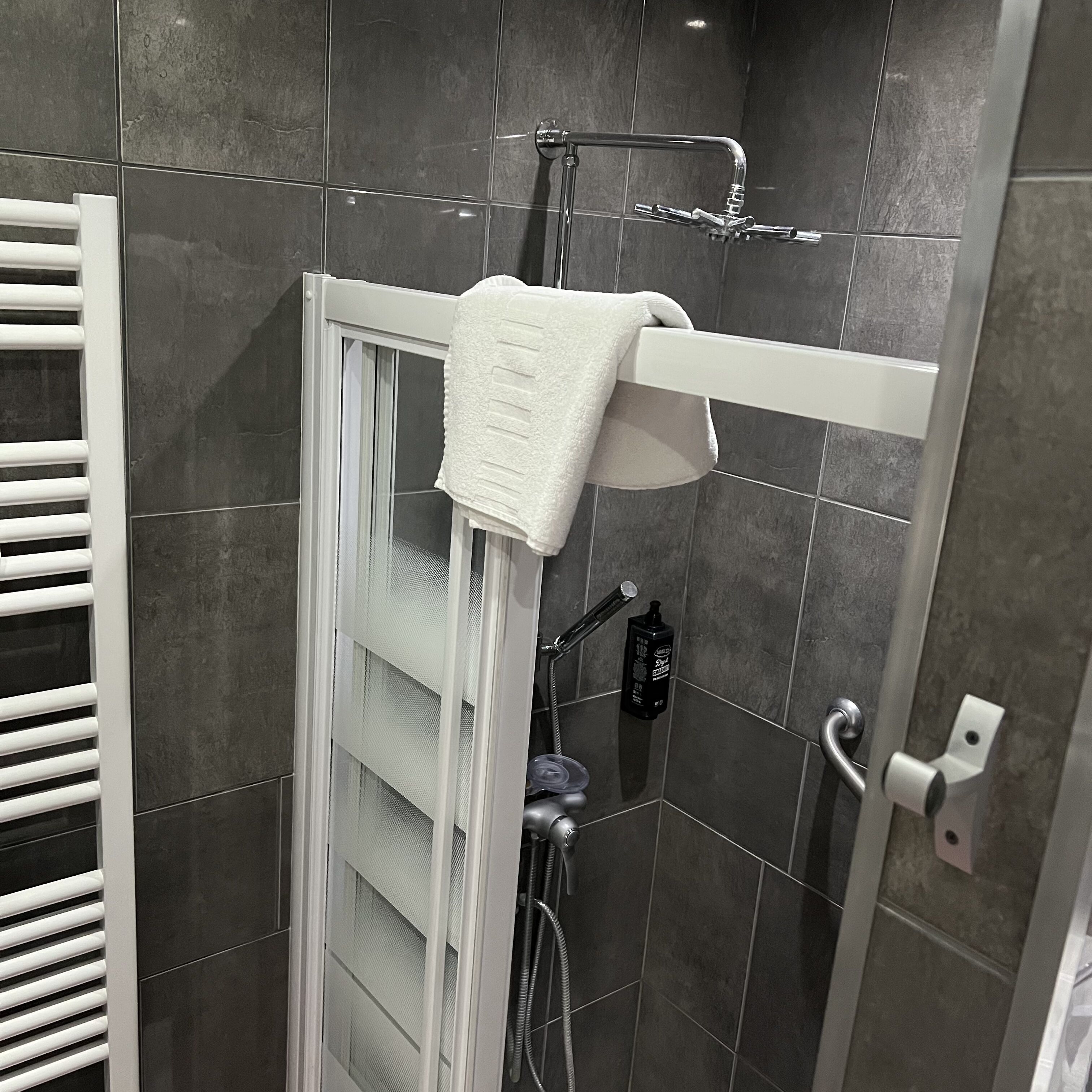 Una ducha con una toalla blanca colgada en un perchero, un cabezal de ducha manual y una botella de champú en la pared.