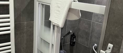 Une douche avec une serviette blanche suspendue à un porte-serviettes, une pomme de douche à main et une bouteille de shampoing fixée au mur.