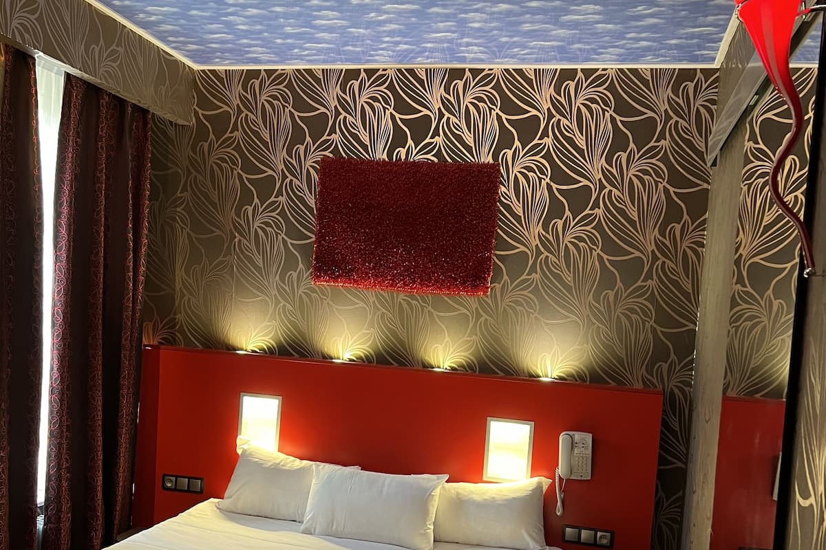 A hotel room with a bed, a red headboard, a patterned wall, and a ceiling with a cloud design.
