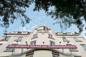 The facade of Le Grand Hotel de Valencia with a prominent sign, multiple windows, and a flag. - Le Grand Hôtel de Valenciennes (Valenciennes)