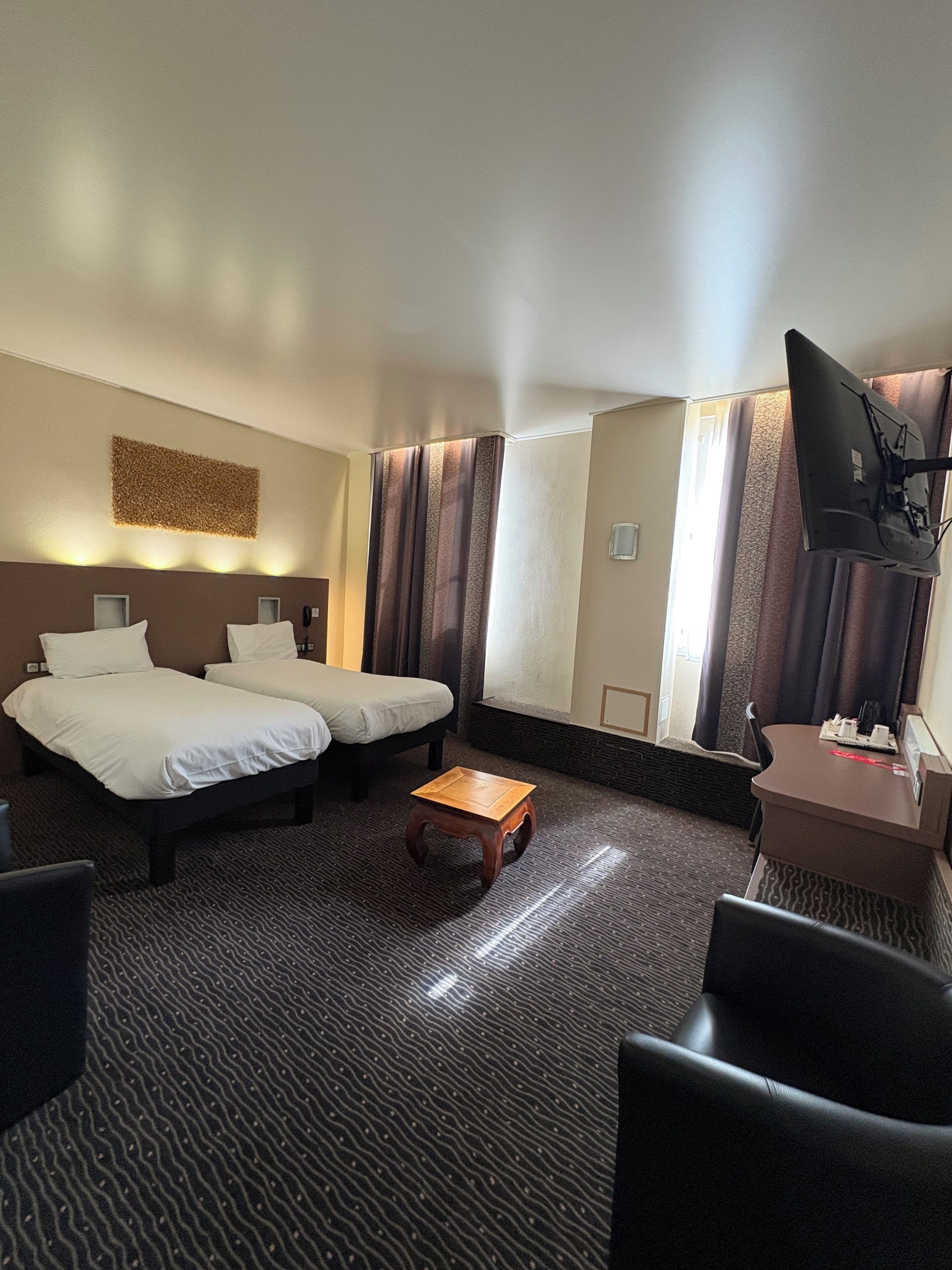 Ein Hotelzimmer mit zwei Betten, einem kleinen Holztisch, einem Schreibtisch mit Stuhl, einem Flachbildfernseher und einer Wanddekoration.