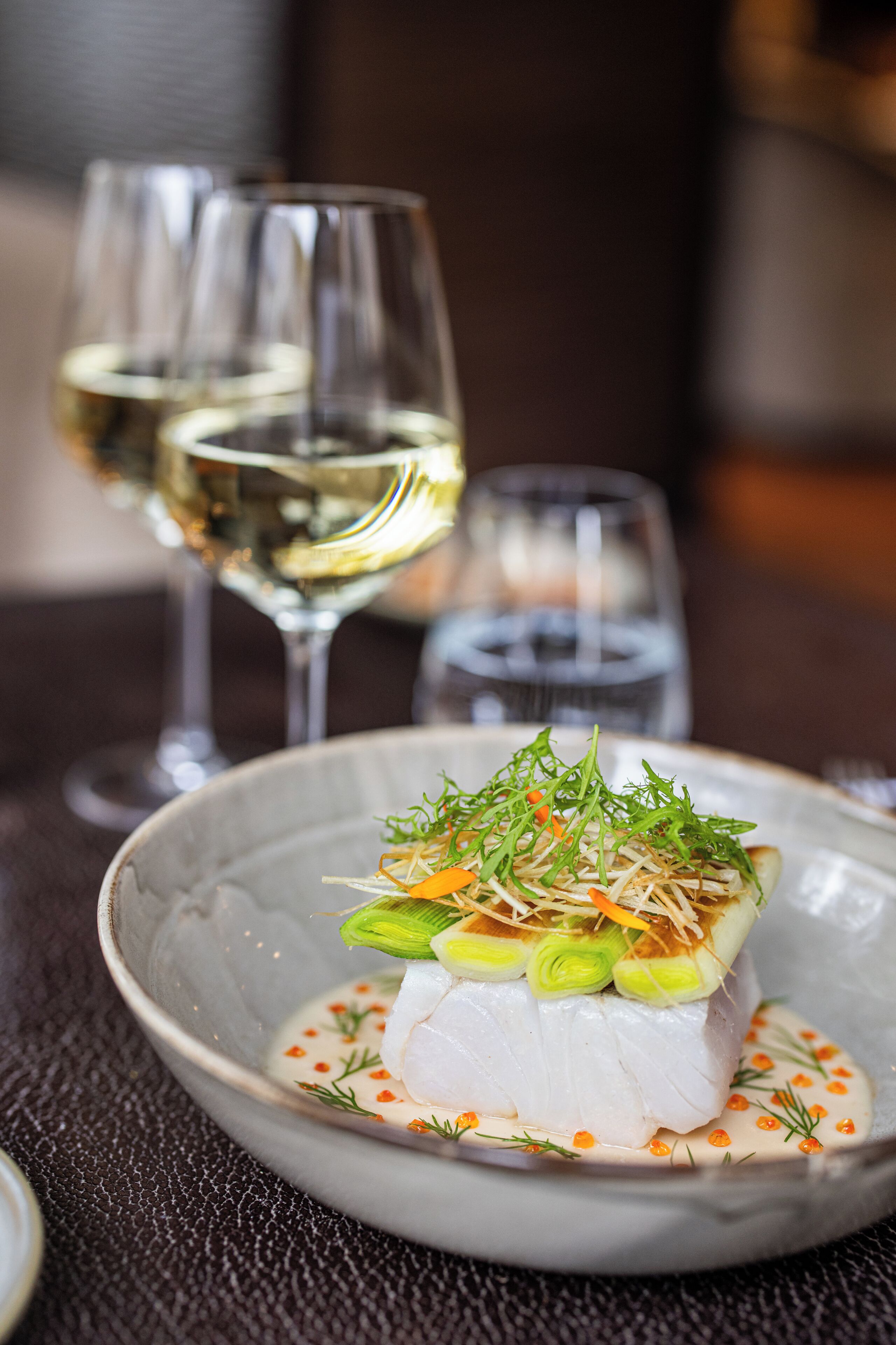A dish with white fish, green onions, and carrot garnish on a plate, accompanied by two glasses of white wine.