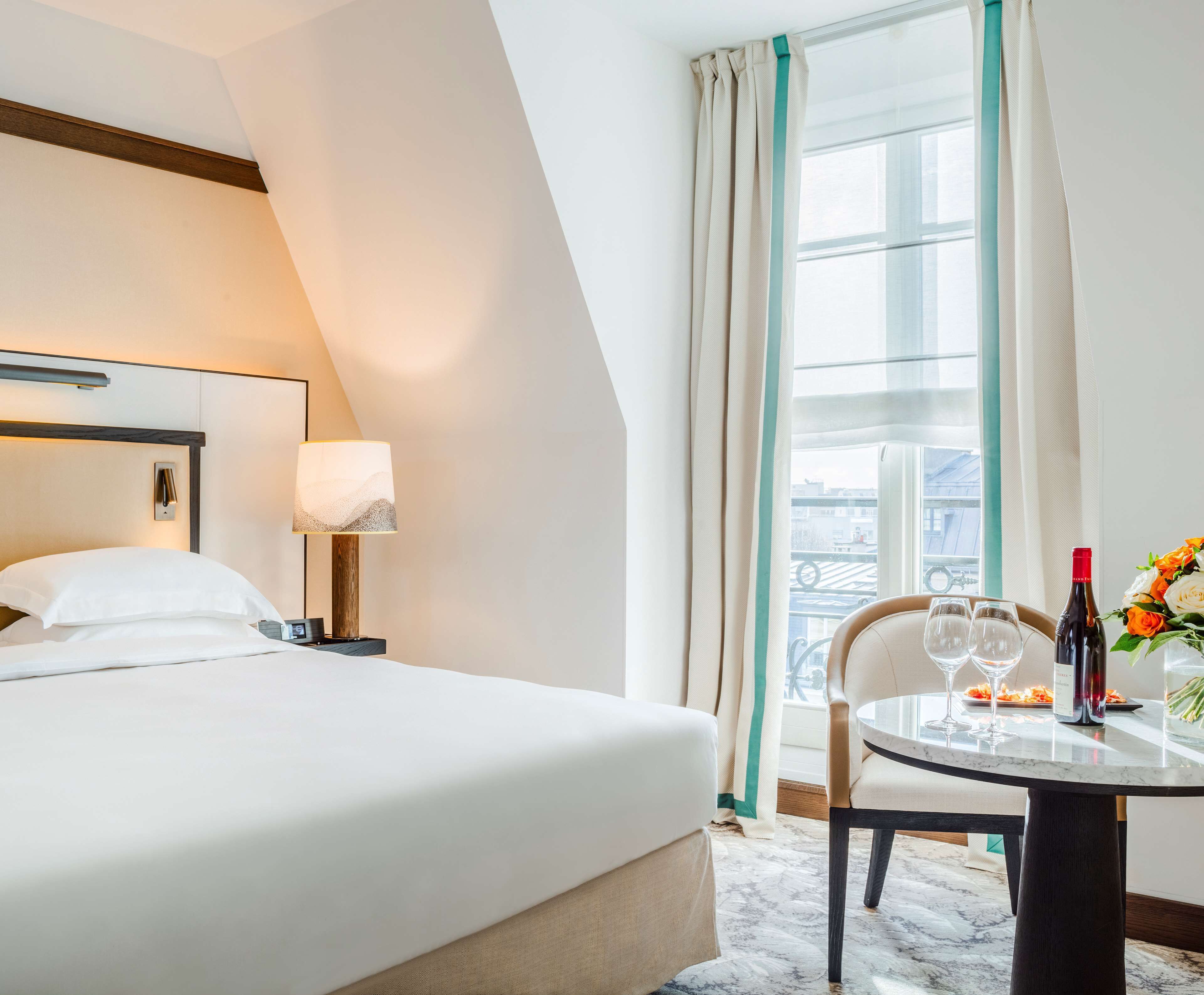 A hotel room with a bed, a bedside table with a lamp, a round table with wine glasses and a bottle, and a view of the city through a large window.