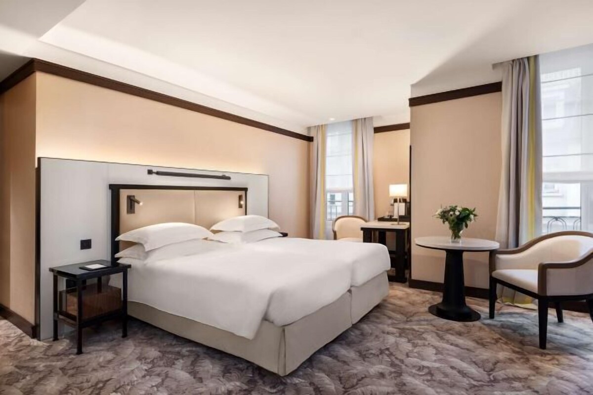 A hotel room with a large bed, a bedside table, a chair, a small table with a lamp, and a window with curtains.