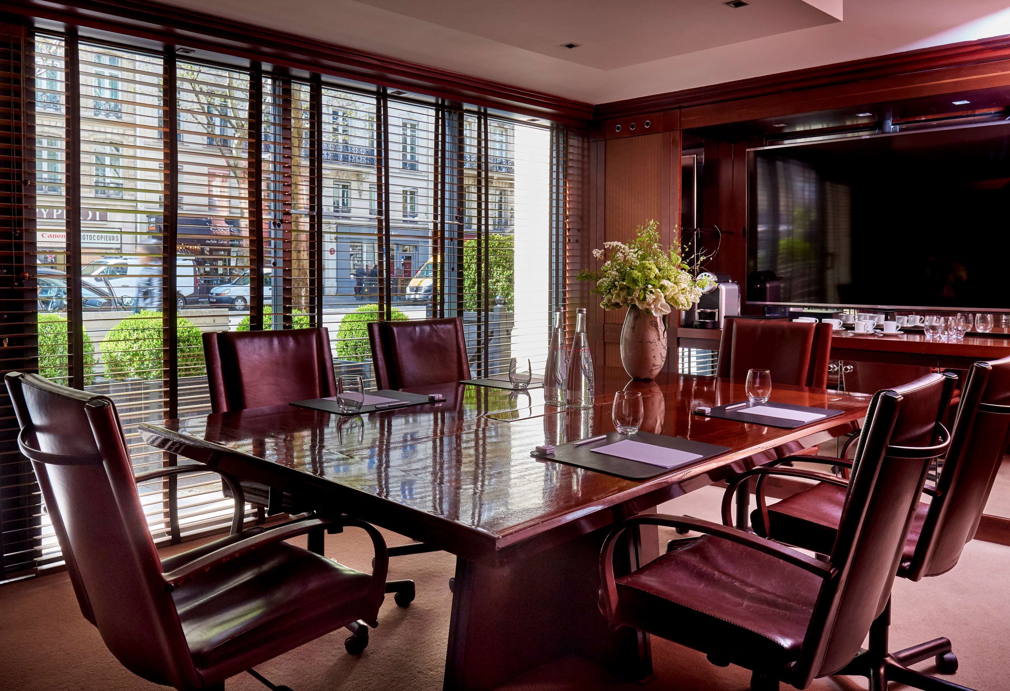A conference room with a glass table, leather chairs, a vase with flowers, and a large window with blinds.