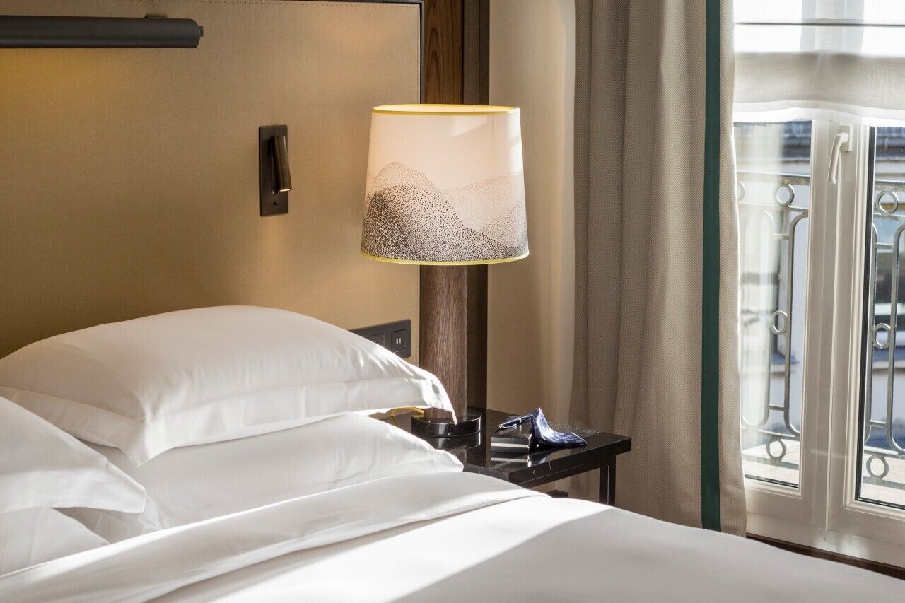 A hotel room with a bed, a nightstand, a lamp, and a window with a view.